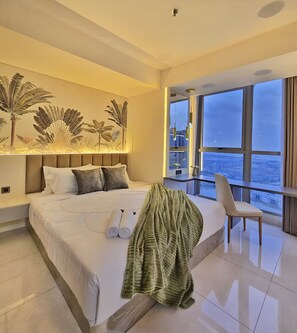 A modern hotel room with a large bed, a balcony with a city view, and a decorative wall with plant art. - Staysanliving Goldcoast PIK (Jakarta)