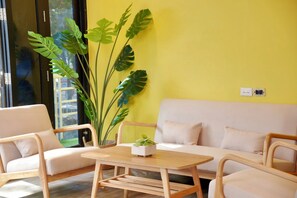 A modern living room with a yellow wall, a white sofa, wooden chairs, a wooden table, and a large plant. - Bed & Beach Hostel Karon (Karon)