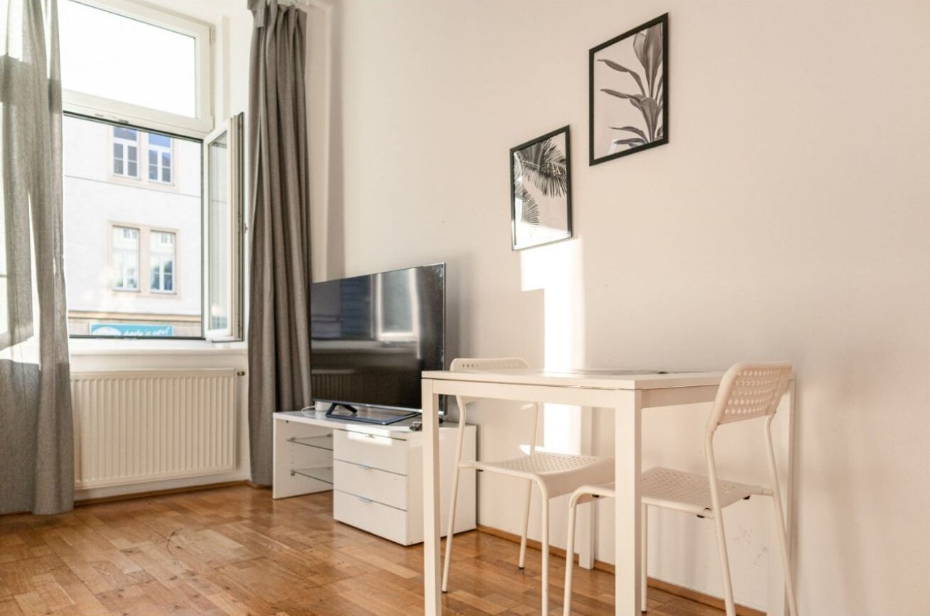 Ein Zimmer mit Fernseher, einem kleinen weißen Tisch, einem Stuhl, einem Fenster mit Vorhängen und zwei gerahmten Bildern an der Wand.