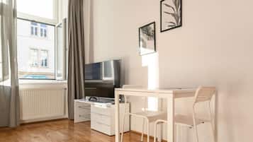 Ein Zimmer mit Fernseher, einem kleinen weißen Tisch, einem Stuhl, einem Fenster mit Vorhängen und zwei gerahmten Bildern an der Wand.