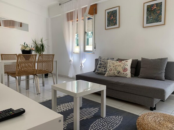 A modern living room with a grey sofa, white coffee table, and wicker chairs. There are two framed pictures on the wall, a potted plant, and a window with sheer curtains.