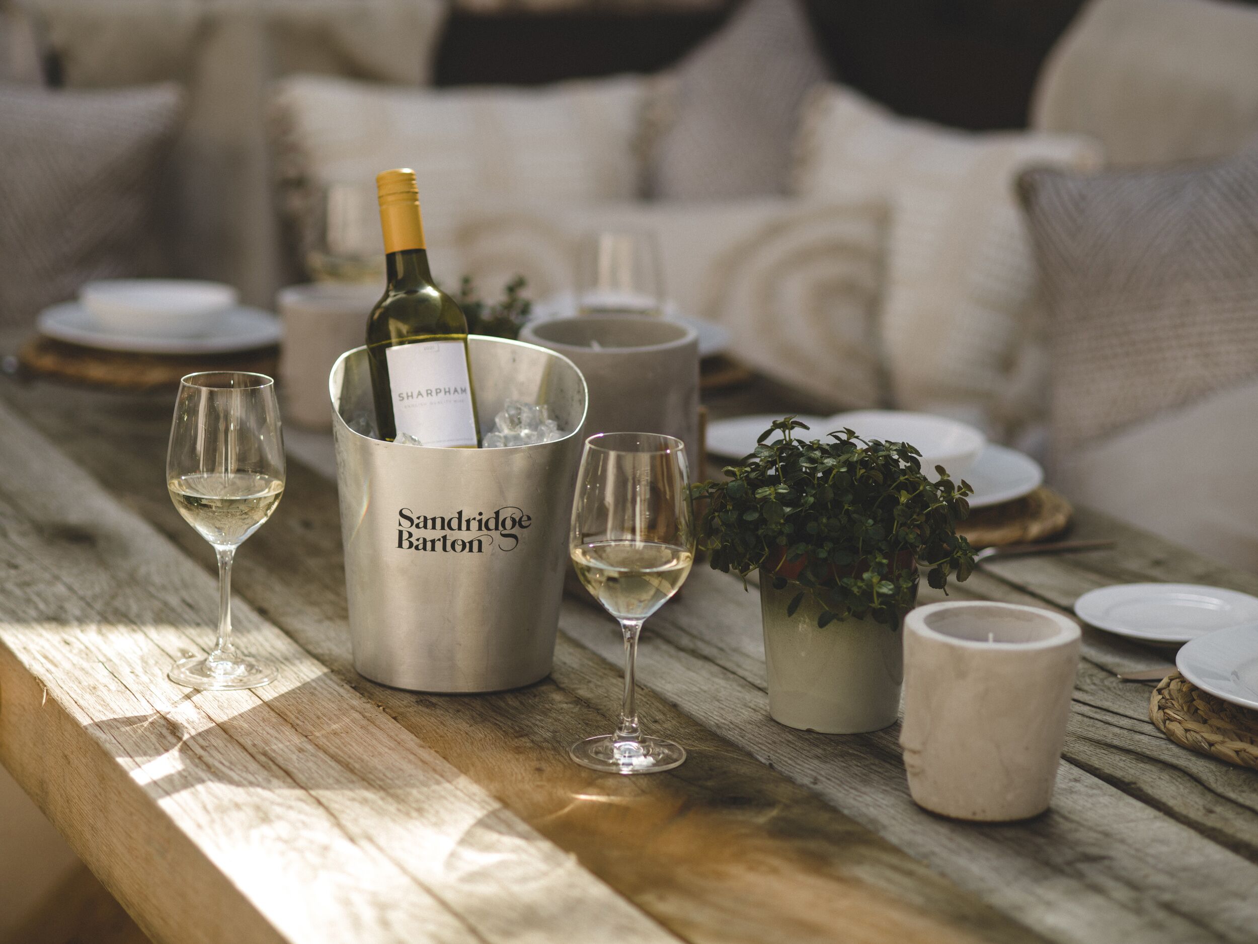Une table en bois sur laquelle se trouve un récipient en métal portant l’inscription « Sandridge Barton ». Ce récipient contient une bouteille de vin, deux verres à vin et une plante en pot.