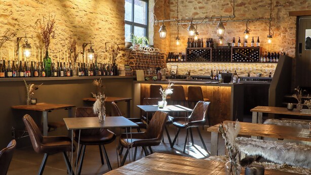 A rustic bar area with wooden tables and chairs, a stone wall, and a variety of wine bottles and glasses. - Lower Well Farm House (Totnes)