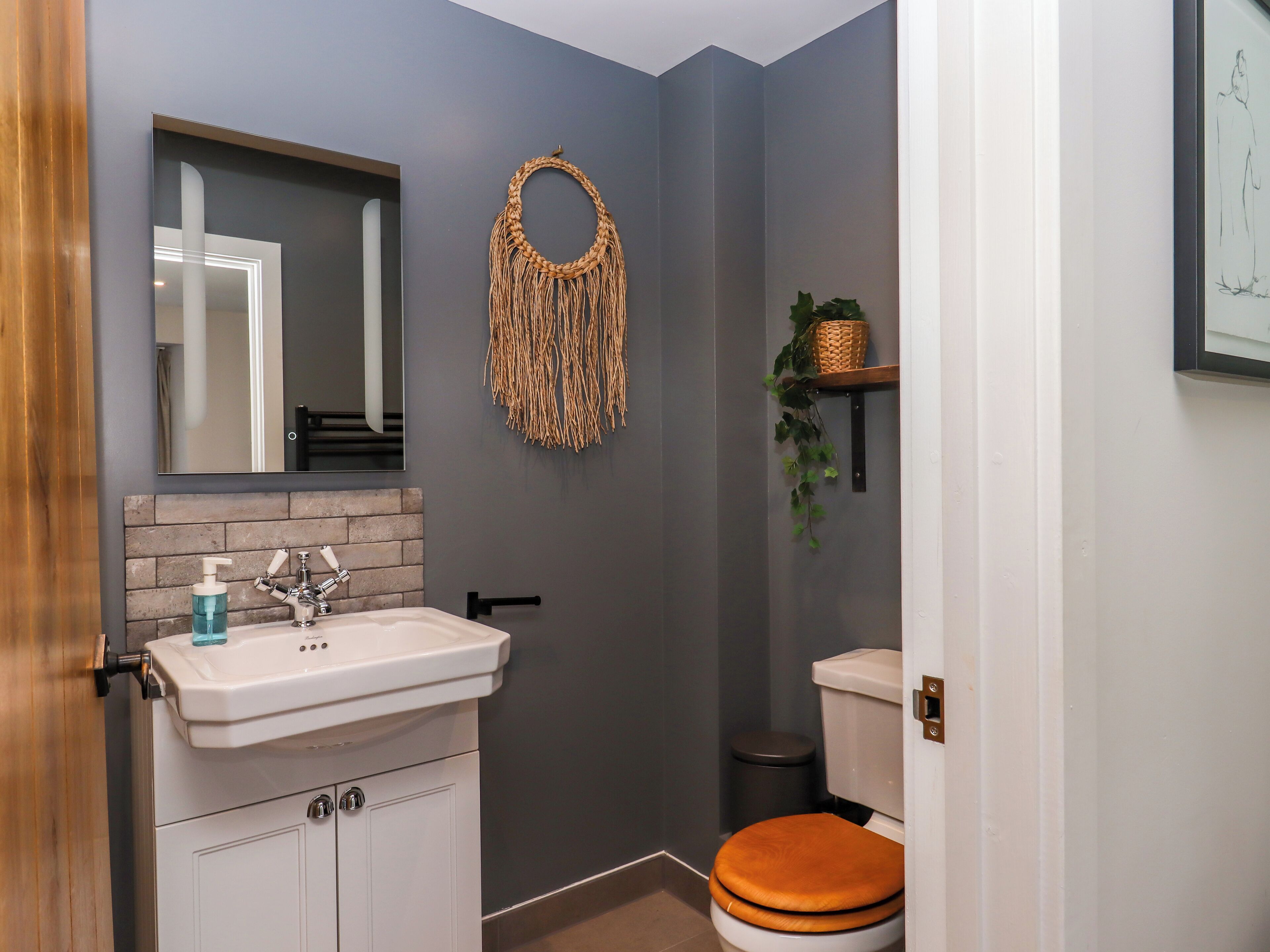 A bathroom with a white sink, a mirror, a brick-patterned wall, a toilet with an orange seat, and a wall-mounted decorative item.