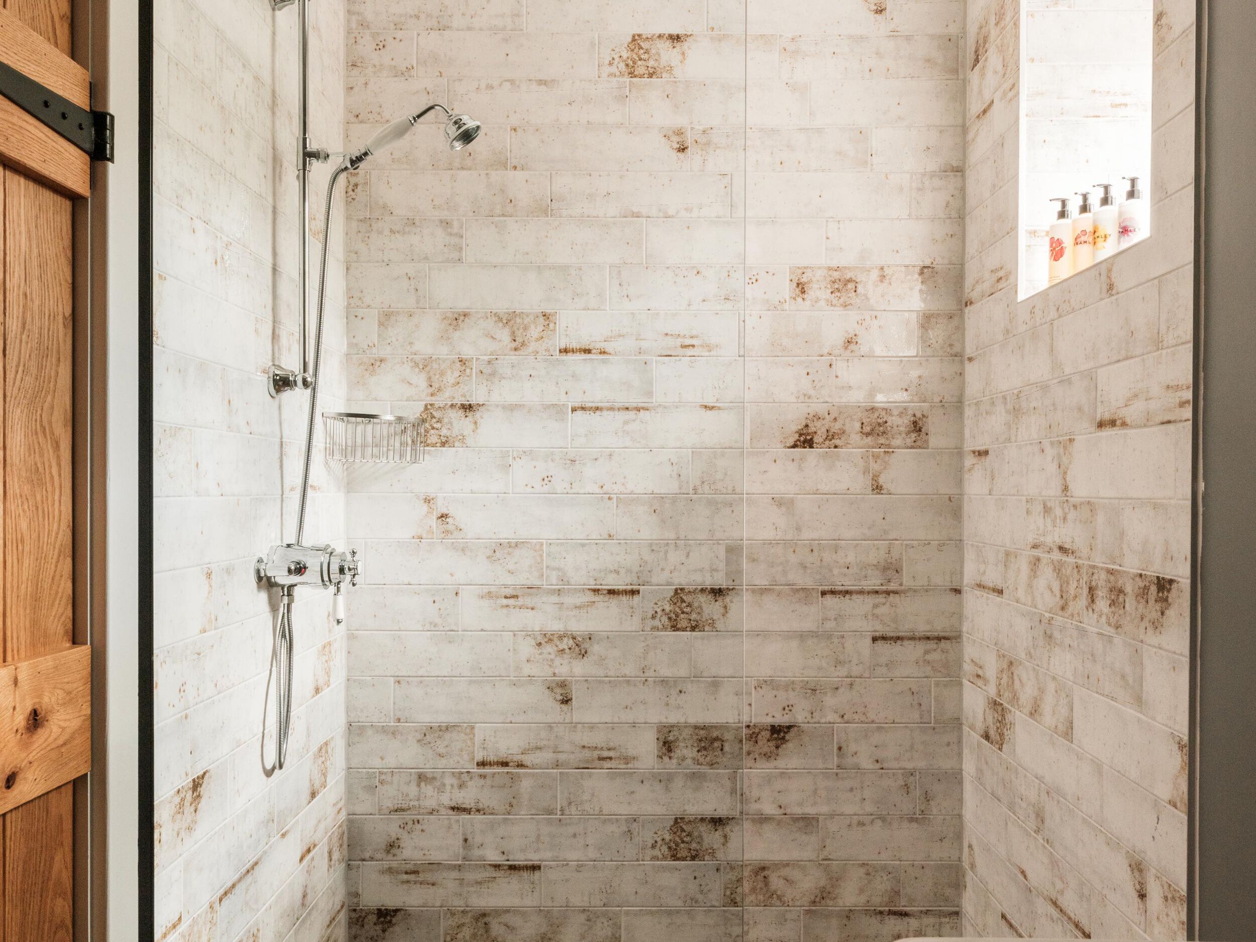 A shower with a wooden door, white tiled walls, and a visible window with bottles.