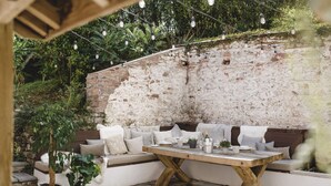 An outdoor seating area with a wooden table, cushioned benches, and string lights. - Lower Well Farm House (Totnes)