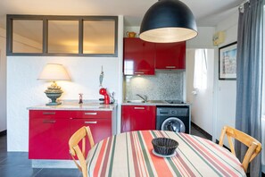Une cuisine compacte avec des meubles rouges, un lave-linge, une table à manger recouverte d’une nappe à rayures et une lampe suspendue.