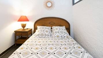 A bed with patterned bedding, a bedside lamp, a small wooden nightstand, and a wall-mounted mirror.