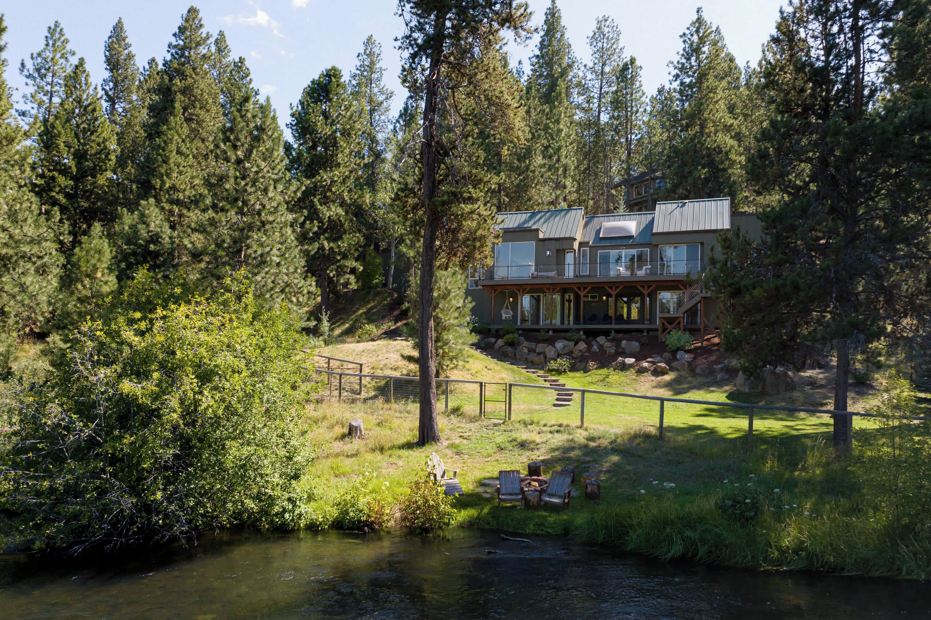 A two-story cabin with a deck, surrounded by dense forest and a stream.
