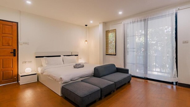A modern bedroom with a bed, grey sofa, and a large window with sheer curtains. - Modern Family House - 4 Bedrooms 5 Mins From Old Town With Gallery - Deja Home (Chiang Mai)