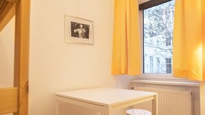 Une pièce avec une table blanche, un petit tabouret, un tableau encadré accroché au mur et une fenêtre ornée de rideaux jaunes.