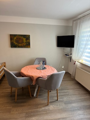 Pieni huone, jossa on pyöreä pöytä, kaksi tuolia, taulu, televisio ja ikkuna, jossa on verhot.