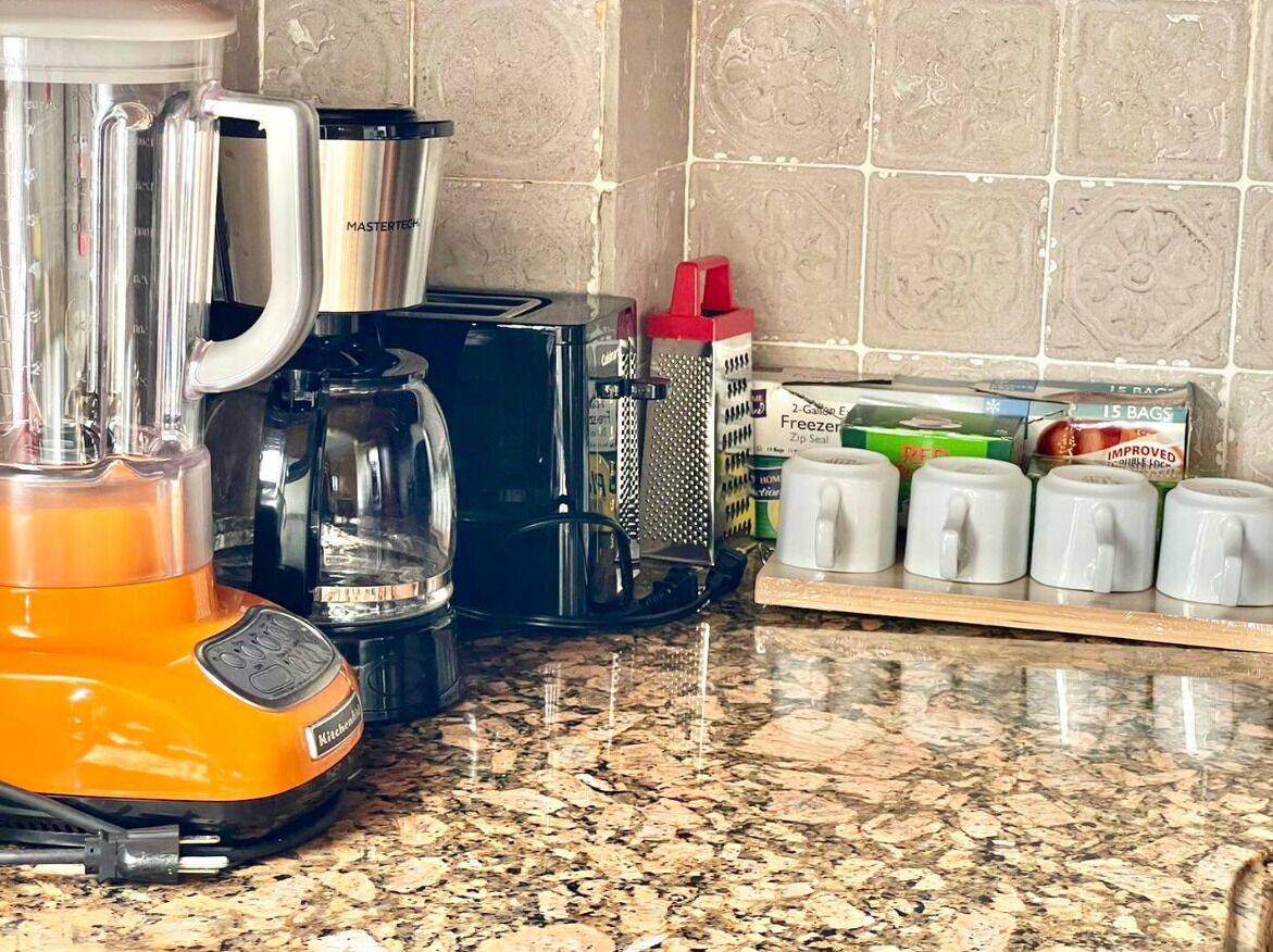 A kitchen counter with a blender, a coffee maker, a grater, and a shelf with mugs.