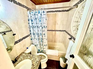 Casa de banho com banheira, duas sanitas, lavatório e cortina de chuveiro com motivos náuticos.
