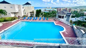 Una piscina con acqua azzurra, circondata da un pavimento in mattoni rossi e ringhiere bianche. Lungo il bordo della piscina ci sono sedie a sdraio blu.