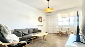Een moderne woonkamer met een grijze bank, een houten stoel, een klein rond tafeltje en een televisie op een witte kast.