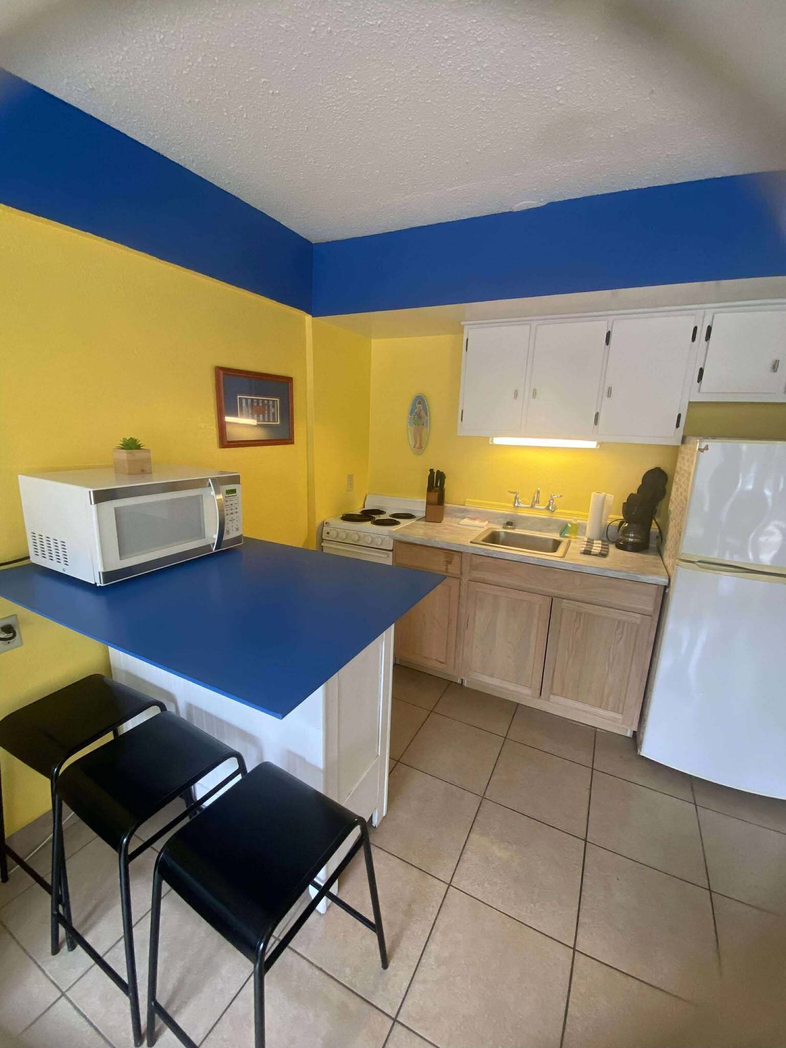 A compact kitchen with a blue countertop, white cabinets, a microwave, a stove, and a refrigerator. There are two black stools and a small potted plant on the countertop.