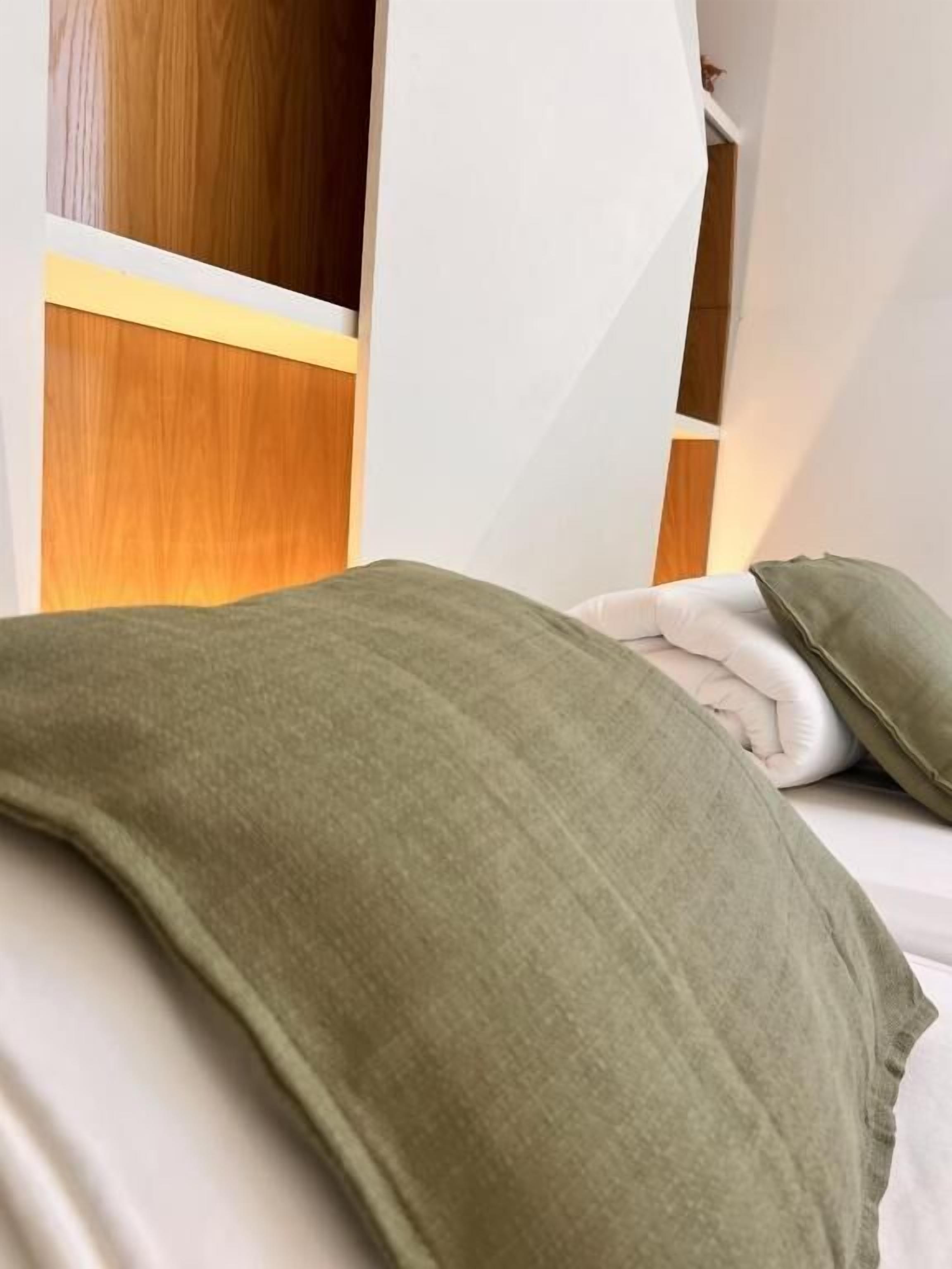 A bed with a green blanket and white pillows, wooden headboard, and white walls.
