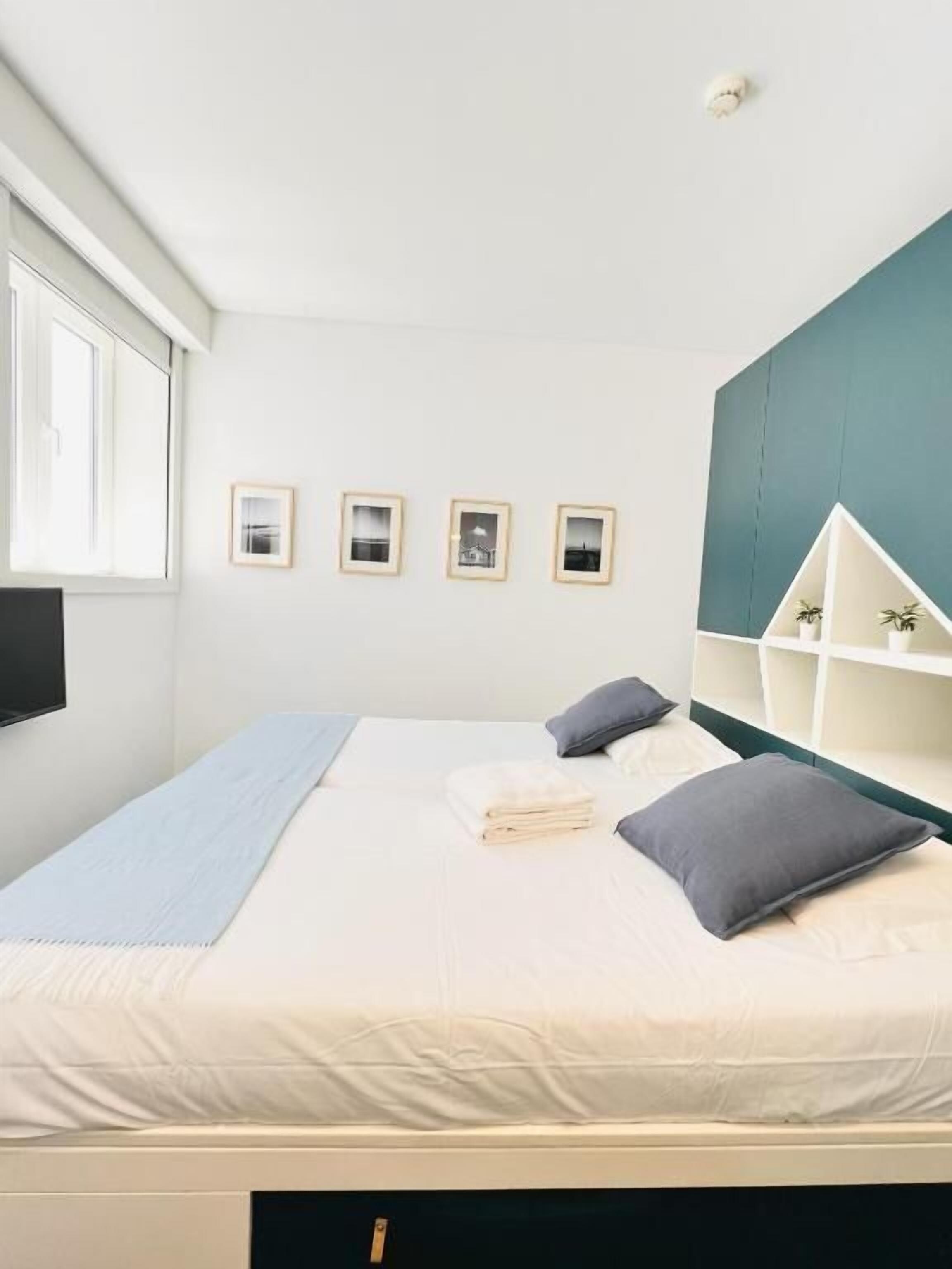 Un dormitorio moderno con cama, almohadas, mesita de noche, ventana y cuadros en la pared.
