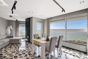 A modern dining area with a white table, grey chairs, and a view of the sea.