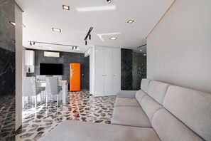 A modern living room with a sectional sofa, a dining area with a white table and chairs, and a kitchen with a black backsplash.