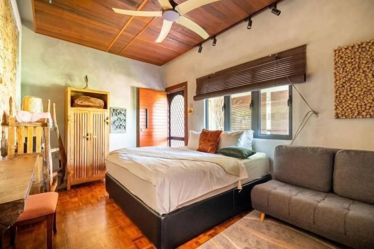 A bedroom with a wooden ceiling, a bed with white bedding, a gray sofa, and a wooden cabinet.