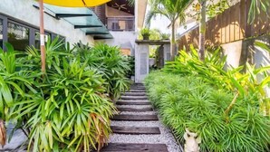 Un sendero estrecho bordeado por plantas verdes exuberantes, que conduce a un edificio de diseño moderno y una sombrilla amarilla.
