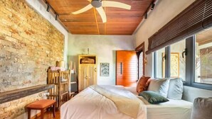 Un dormitorio con pared de ladrillo, ventilador de techo, cama con almohadas, banco y ventana.