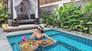 Un área de piscina con terraza de madera, zona de estar cubierta y abundante vegetación.