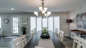 Uma sala de jantar com uma mesa comprida, cadeiras brancas, um candelabro e um relógio na parede.