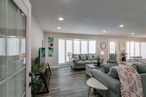 A spacious living room with a grey sofa, a coffee table, and a dining area with a wooden table and chairs. There is a large window with blinds, a clock on the wall, and a painting on the wall. - Residence (1335 Whitt Ln) (Clarksville)