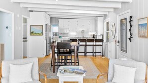 A spacious living room with a dining area, a kitchen with white cabinetry, and a large window. - Walk to beach access - hot tub, dog friendly, sleeps 9! (Oak Island)