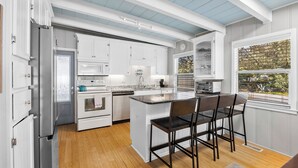 A kitchen with white cabinets, a central island with bar stools, a refrigerator, and a window with a view of greenery. - Walk to beach access - hot tub, dog friendly, sleeps 9! (Oak Island)
