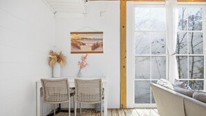 A cozy room with a wooden floor, a white wall, a framed painting, a vase with dried plants, and a window with a view of trees. - Walk to beach access - hot tub, dog friendly, sleeps 9! (Oak Island)