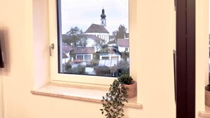 A window with a view of a residential area, including houses and a church steeple. - Modern apartment with kitchen & parking (Stephansposching)