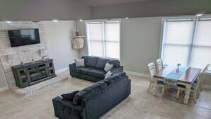 A living room with a grey sofa, a TV mounted on a brick wall, a dining table with chairs, and large windows. - Magnolia Summary:<br>Hil (Clarksville)