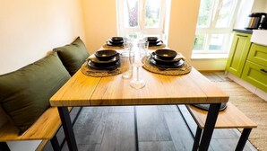 Ett matbord dukat med svarta tallrikar, klara glas och ett vinglas, placerat i ett rum med en grön soffa, ett fönster med persienner och en köksdel med gröna skåp.