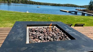 A square fire pit with rocks and embers, surrounded by a wooden deck, overlooking a lake with a dock and a bridge. - 7-bed lakefront house with sauna, beach & boat launch — 15 min from Thunder Bay. (Thunder Bay)
