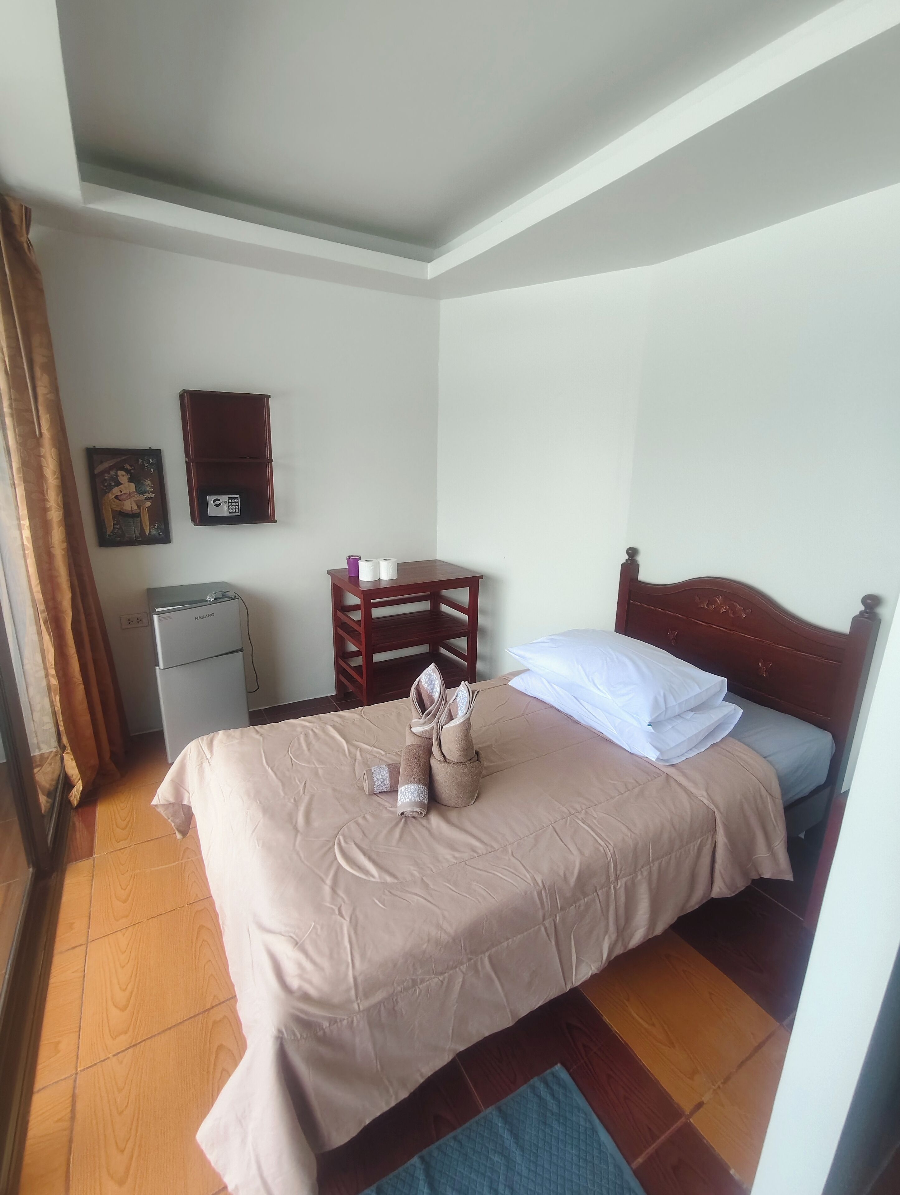 Ein ordentlich bezogenes Bett mit einer braunen Tagesdecke, ein Nachttisch aus Holz mit einer Lampe, ein kleiner Tisch mit einem Stuhl und ein wandmontierter Schrank.