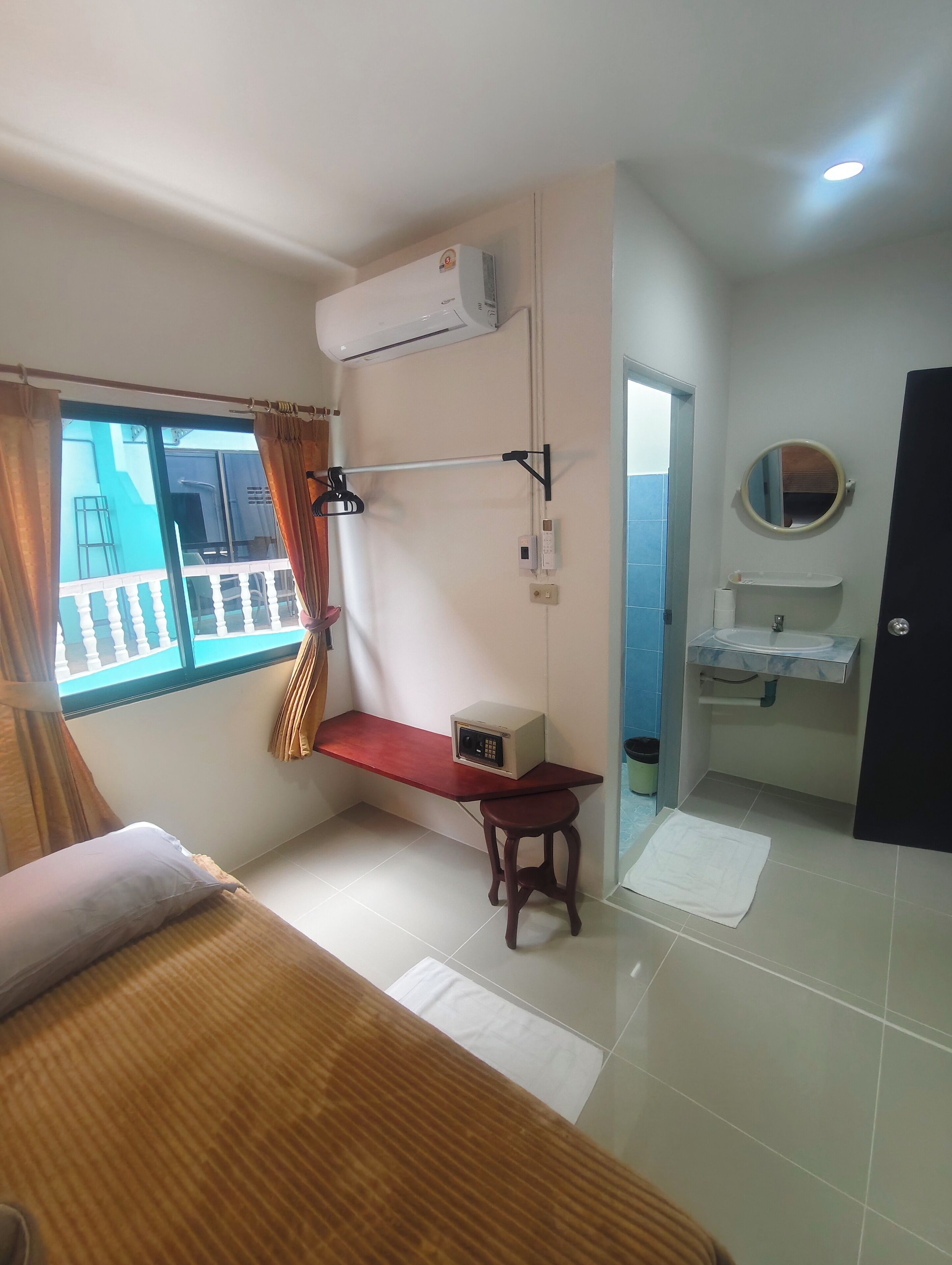 A compact hotel room with a bunk bed, a small desk with a heater, a window with curtains, a bathroom with a sink and mirror, and a view of the outside balcony.
