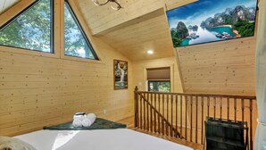 A wooden cabin room with a bed, a painting, a TV, and a balcony with a railing. - The Hocking Hills Treehouse (Rockbridge)