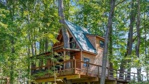 A wooden treehouse with a metal roof, built on stilts among tall trees. - The Hocking Hills Treehouse (Rockbridge)