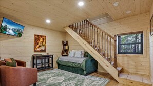 A cozy room with a wooden staircase, a bed, a sofa, a TV, and a bookshelf. - The Hocking Hills Treehouse (Rockbridge)