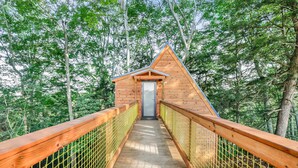 A wooden cabin with a triangular roof, accessed by a wooden bridge with a netted railing, surrounded by dense forest. - Pikachu's Palace Treehouse (Rockbridge)