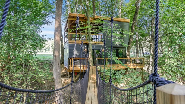 A wooden suspension bridge leading to a multi-level wooden cabin surrounded by trees. - The Country Club Treehouse (Rockbridge)