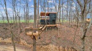A modern cabin with a wooden deck and a balcony, surrounded by trees and a dirt path. - The Country Club Treehouse (Rockbridge)