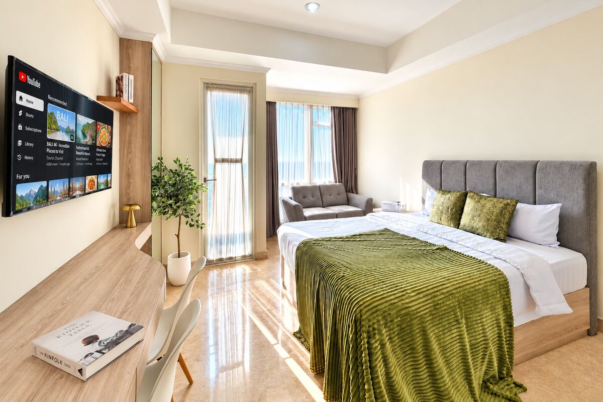 A modern hotel room with a bed, a seating area, a TV displaying a travel channel, and a potted plant.