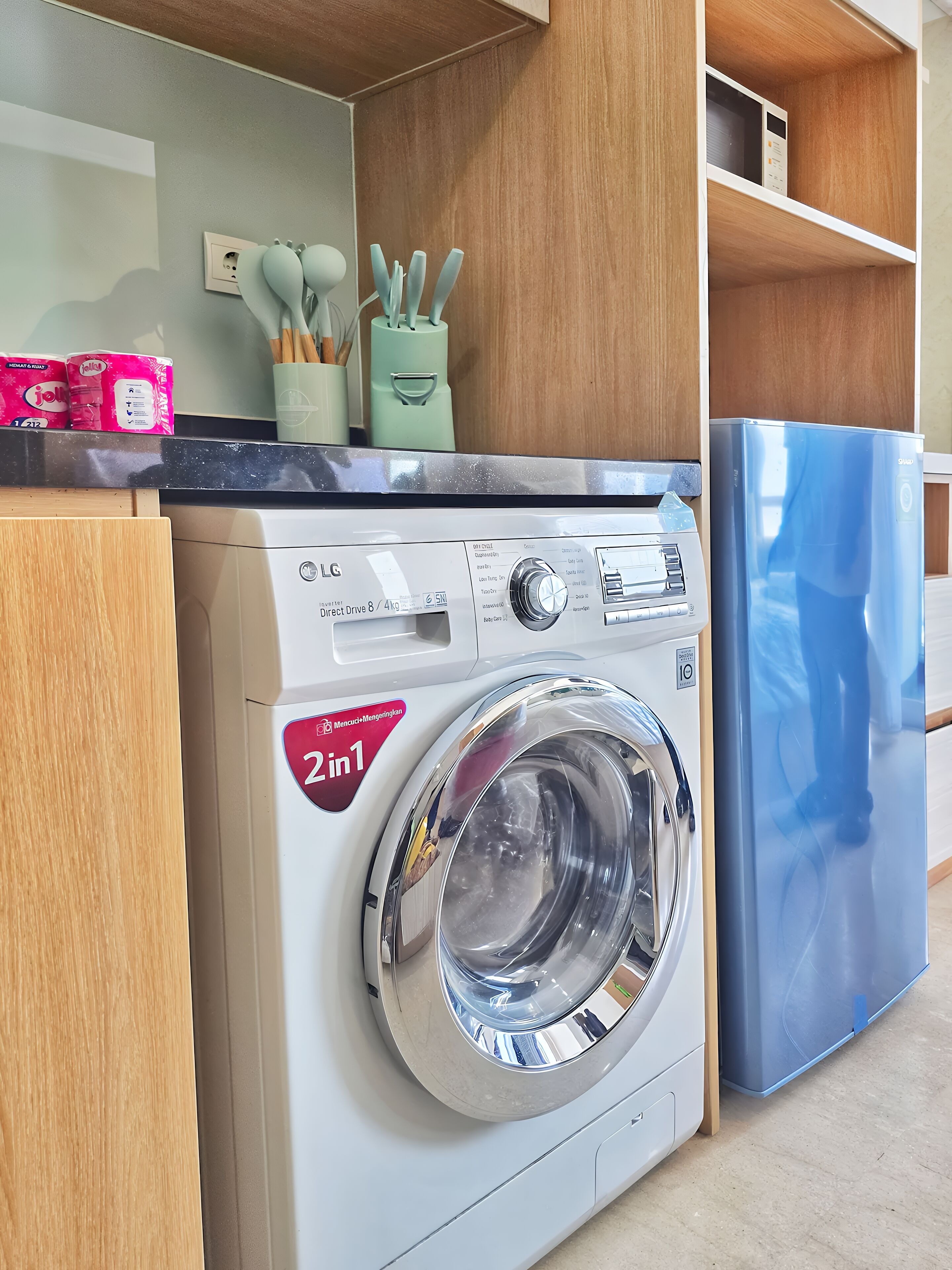 A compact washer and dryer unit with a 2 in 1 feature, placed in a modern kitchen with wooden cabinets and a blue refrigerator.