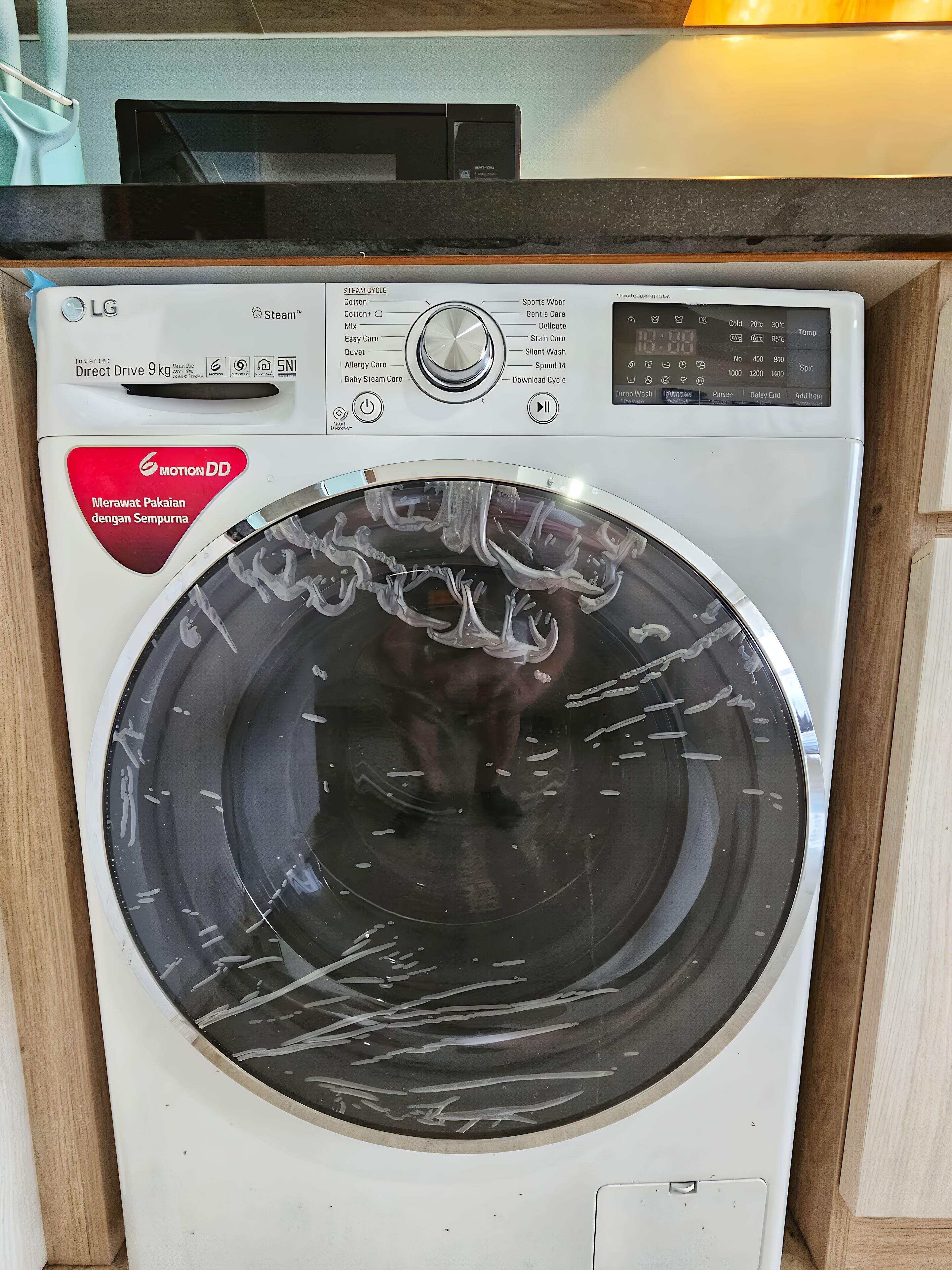 A front view of a washing machine with a control panel and a red label indicating 'Motion DD' and 'Merawat Pakaian dengan Sempurna'.