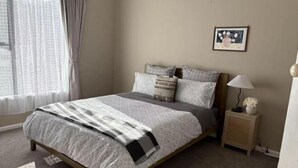 A bedroom with a bed, a bedside table, a lamp, a picture on the wall, and a window with curtains. - The Nest on Till (Oamaru)
